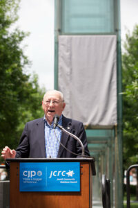 Israel Arbeiter speaks to the crowd at the 2017 Rededication Ceremony of the New England Holocaust Memorial in Boston. He is standing in front of a glass panel about to be unveiled that had to be replaced after it was vandalized. That panel contained the number that is tattooed on his arm. Photo/Courtesy of CJP