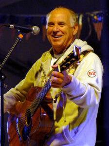 Jimmy Buffett performing on stage with a guitar, smiling, wearing a light hoodie, and engaging with the audience, reflecting the lively spirit of his music inspired by experiences like his "Boat Drinks" story.