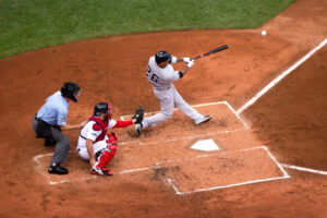 Baseball player swinging bat at home plate during a game, with catcher in red gear and umpire in blue uniform, representing the Boston Red Sox versus Detroit Tigers on Patriots' Day.