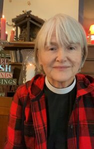 Rev. Julia Dunbar, Episcopalian priest in plaid shirt, smiling in front of a bookshelf, reflecting her journey in pastoral care and retirement.